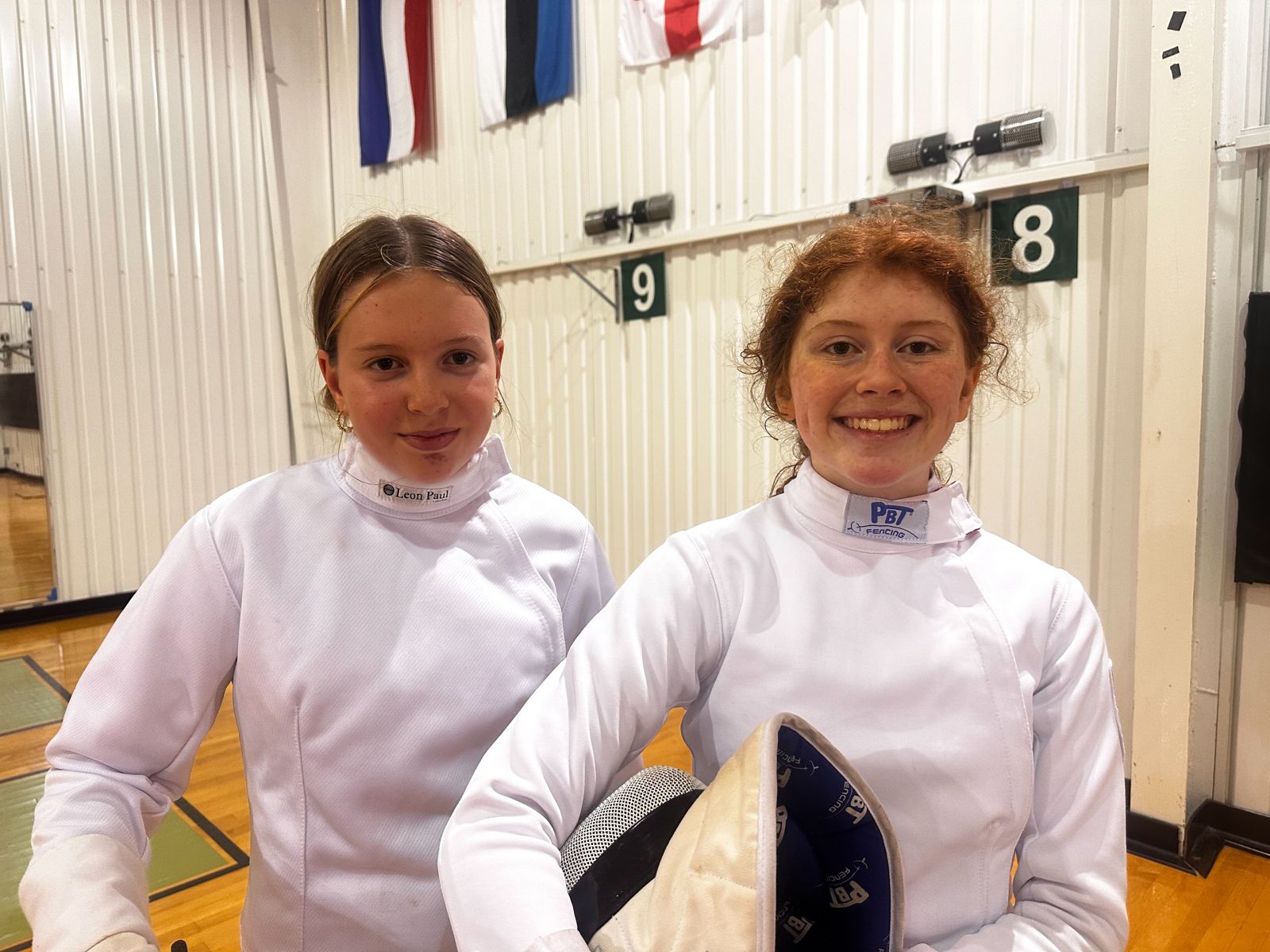 Two young fencers in white fencing jackets smiling at the academy, one holding her mask